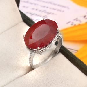 Può includere: Un anello d'argento con un grande rubino ovale. Il rubino è di un rosso intenso ed è circondato da un alone di pietre più piccole e trasparenti. L'anello è presentato in una scatola foderata di velluto.