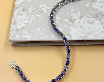 May include: A silver bracelet with oval, dark blue gemstones. The bracelet is laid out on a light beige surface. The clasp is visible at the end of the bracelet. The gemstones are set closely together.