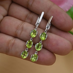 May include: A pair of silver dangle earrings featuring three oval-cut, light green peridot gemstones. The earrings have a simple, elegant design, with each gemstone set in a silver bezel. The earrings are shown against a neutral background.