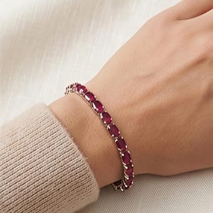 May include: A tennis bracelet featuring a series of oval-cut, deep red rubies set in a polished silver-toned metal band. The stones are arranged in a continuous line, each secured by individual metal prongs, creating a uniform and elegant design.