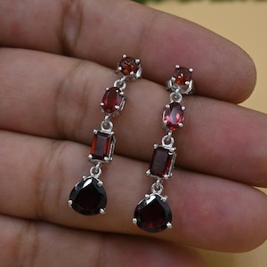 Könnte beinhalten: Ein Paar Ohrhänger mit silberfarbener Fassung. Jeder Ohrring hat eine Reihe roter Edelsteine in verschiedenen Formen: rund, oval und rechteckig. Der größte Edelstein hat eine Tropfenform. Die Ohrringe werden vor einem neutralen Hintergrund gezeigt.