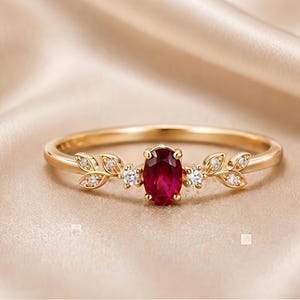May include: A gold ring featuring a central oval-cut deep red gemstone flanked by two small clear round stones on each side. The band is decorated with delicate gold leaf motifs that extend outward from the centre, set with tiny clear stones.
