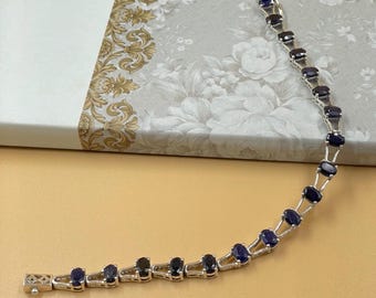 May include: A silver bracelet with oval, dark blue gemstones. The gemstones are set in individual silver links, creating a delicate and elegant design. The bracelet is displayed on a light beige surface, with a floral patterned background.