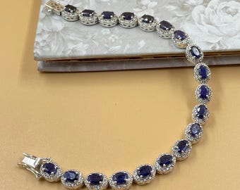 May include: A silver bracelet featuring oval, dark blue gemstones surrounded by small, clear stones. The bracelet is laid out on a light-colored surface, showcasing its elegant design and clasp.