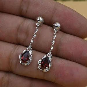 May include: A pair of silver drop earrings. Each earring has a teardrop-shaped, deep red gemstone set in a decorative silver frame. The gemstones are suspended from a twisted silver chain.