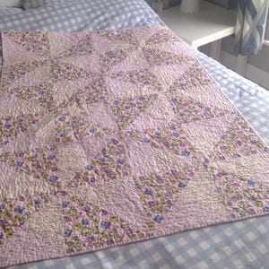 May include: A pink and floral quilt with a patchwork design, resting on a bed with a blue and white checkered pattern. The quilt features a repeating pattern of floral squares in shades of pink, purple, and green.