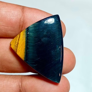 Könnte beinhalten: Ein polierter, dreieckiger Edelstein mit einem auffälligen Farbkontrast. Der Stein weist einen tiefen Blaugrün-Ton mit einem leuchtenden Gelbgold-Bereich auf einer Seite auf. Die polierte Oberfläche reflektiert das Licht und hebt die natürlichen Muster hervor.