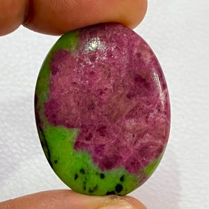 Könnte beinhalten: Ovaler Edelstein mit einer lebendigen Farbpalette. Der Stein zeigt eine auffällige Kombination aus tiefem Magenta und leuchtendem Grün. Das Magenta dominiert die Mitte, während das Grün einen Rand mit schwarzen Sprenkeln bildet. Der Stein wird von einem Finger gehalten.