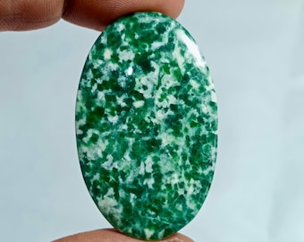 May include: An oval gemstone, predominantly green with white flecks, held between fingers. The polished surface of the stone reflects light, showcasing its natural patterns. The gemstone's shape is approximately 3 cm in length.