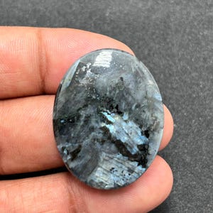 May include: An oval-shaped labradorite gemstone featuring a polished surface with a dark grey base colour and iridescent blue flashes. The stone displays a complex internal structure with visible crystalline patterns and dark inclusions throughout.