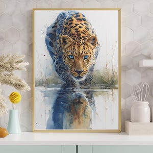 Puede incluir: Una impresión en acuarela enmarcada de un leopardo con ojos azules, caminando hacia el espectador. El pelaje del leopardo es una mezcla de manchas marrones y negras, con un fondo azul y blanco. La impresión se muestra sobre una superficie blanca.