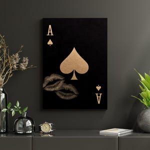Op de afbeelding: Zwart canvas kunstwerk met het ontwerp van de Aas van Schoppen speelkaart. De kaart heeft gouden accenten, waaronder het schoppensymbool, de letter "A" en een paar lippen. Het kunstwerk wordt getoond op een donkergrijze muur.