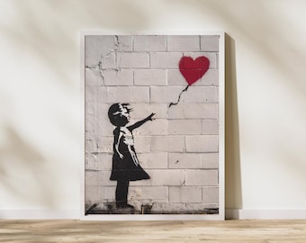 Puede incluir: Obra de arte enmarcada que representa una silueta negra de un niño que alcanza un globo rojo en forma de corazón. La obra es un esténcil sobre una pared de ladrillos blancos, con un marco blanco y un suelo de madera.