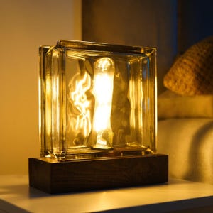 Può includere: Lampada a blocchi di vetro trasparente con una lampadina visibile all'interno, posta su una base di legno marrone scuro. La lampada emette un bagliore caldo, ideale per un comodino o una scrivania. Il blocco di vetro ha un aspetto strutturato.