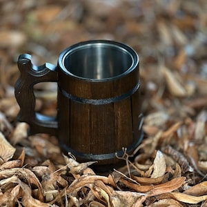 May include: A dark wooden beer mug with a stainless steel interior. The mug has a carved handle and is banded with black trim. The mug is sitting on a bed of dried leaves, creating a rustic, outdoorsy feel.