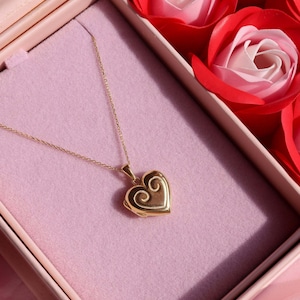 May include: A gold-colored heart-shaped locket necklace with a delicate chain, resting inside a pink box. The locket features an ornate swirl design. Red and pink rose decorations are in the background, creating a romantic theme.