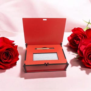 Può includere: Una scatola di legno rossa con coperchio incernierato, aperta per rivelare un piccolo scomparto e un fiammifero. La scatola ha finiture nere. Rose rosse disposte su uno sfondo rosa.