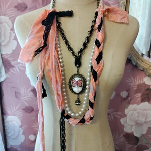 May include: A layered necklace set on a mannequin. The set includes a pearl strand, a chain necklace with a pendant, and a peach-colored fabric scarf with black lace and ribbon accents. The pendant features a butterfly design.