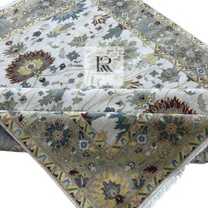Op de afbeelding: Een ingewikkeld patroon vloerkleed met een crèmekleurig middenveld en een rand in tinten grijs, blauw en geel. Het vloerkleed heeft bloemen en geometrische ontwerpen, waardoor een traditionele esthetiek ontstaat. Het vloerkleed is opgevouwen.