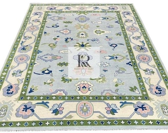 May include: A rectangular area rug with a light blue field and a detailed border. The rug features floral and geometric patterns in shades of green, blue, pink, and cream. The rug has a fringe along the edges.