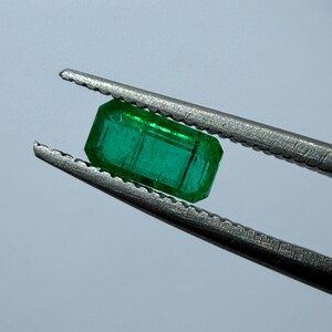 May include: A vibrant, rectangular emerald gemstone, held by silver tweezers. The gem is a deep, transparent green with visible internal features. The tweezers are in sharp focus against a soft, neutral background, highlighting the gem's clarity.