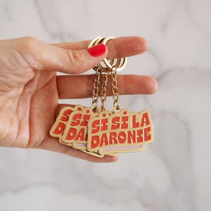 May include: A gold keychain with five red and pink metal tags that say "Sisi La Darone" in a retro font.