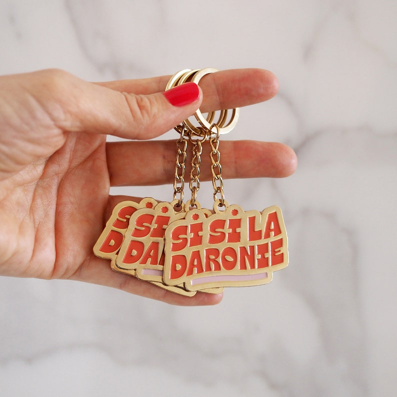 May include: A gold keychain with five red and pink metal tags that say "Sisi La Darone" in a retro font.