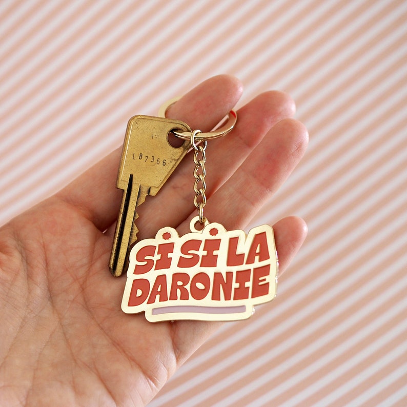 May include: A gold keychain with a red and white enamel charm that says "Sisi La Daronie".