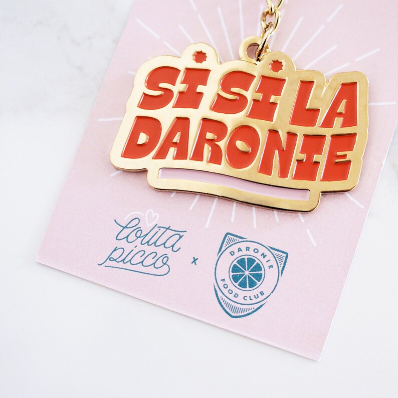 May include: Gold keychain with a red enamel charm that says "Si, Si La Daronie" with a pink and gold background. The keychain is attached to a card with the text "Lolita Picco x Daronie Food Club" in blue and white.