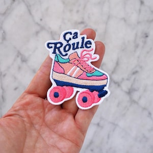 Könnte beinhalten: Gestickter Aufnäher mit einem Retro-Rollschuh-Design in Pink, Koralle, Türkis und Marineblau. Der Aufnäher enthält den französischen Satz "Ca Roule" in einer Schreibschrift, der einen Vintage-Flair verleiht. Der Rollschuh hat rosa Schnürsenkel und Räder.