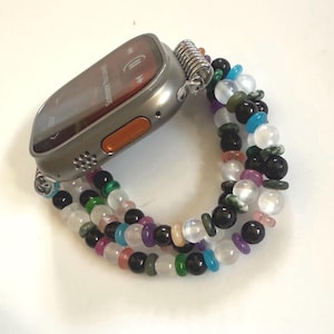 May include: A silver smartwatch with an orange button is attached to a beaded bracelet. The bracelet features a variety of colorful beads, including black, white, green, purple, and pink. The watch screen displays the text "Someone You Loved".