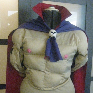 May include: A costume on a mannequin featuring a tan padded torso with pink nipple accents, a dark blue cape, and a red cape. A white skull brooch secures the cape. The costume is displayed on a black dress form.