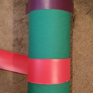 Puede incluir: Un objeto cil&iacute;ndrico verde con una banda roja y otra morada enrolladas alrededor. Las bandas son de un material liso. El objeto est&aacute; sobre una superficie marr&oacute;n.