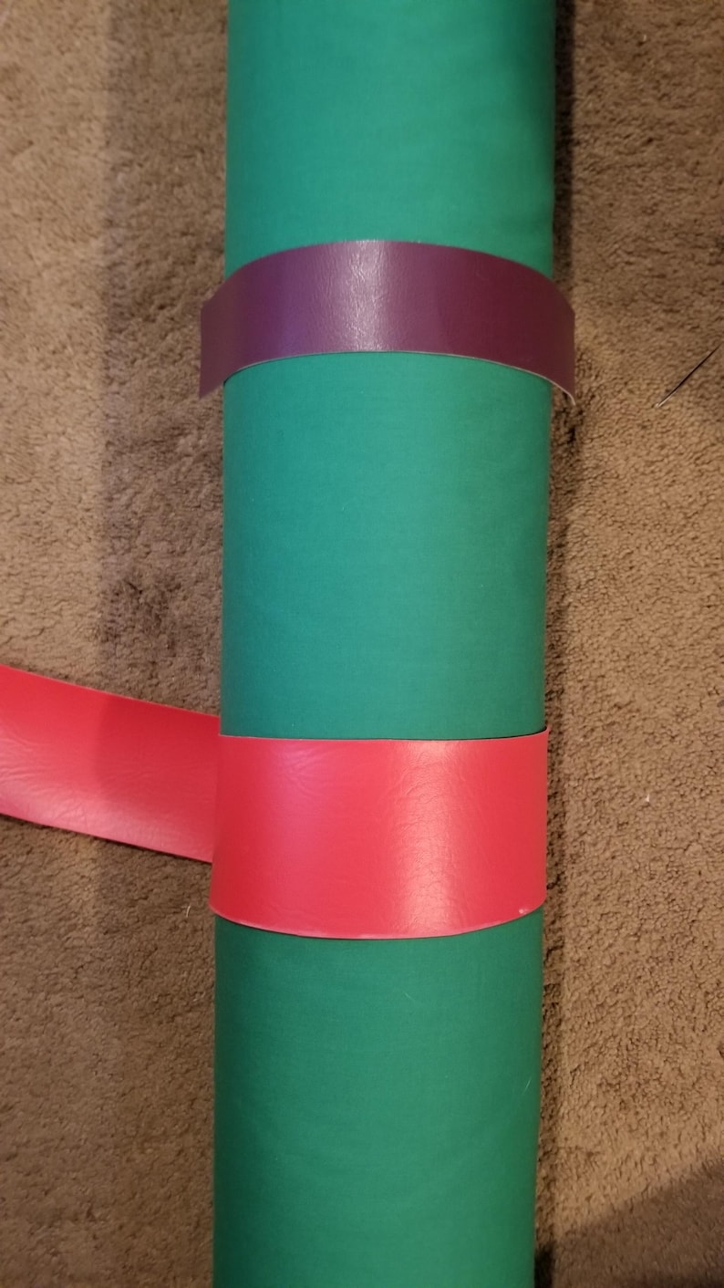 Puede incluir: Un objeto cil&iacute;ndrico verde con una banda roja y otra morada enrolladas alrededor. Las bandas son de un material liso. El objeto est&aacute; sobre una superficie marr&oacute;n.