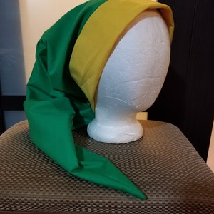 Pode incluir: Um chap&eacute;u verde com uma faixa amarela, com uma ponta longa e pontiaguda. O chap&eacute;u &eacute; exibido em uma cabe&ccedil;a de manequim branca. O chap&eacute;u &eacute; feito de um tecido liso. O fundo &eacute; neutro.