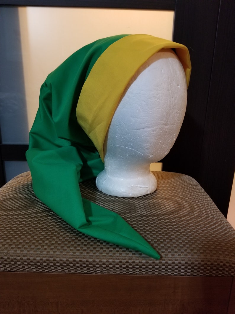 Pode incluir: Um chap&eacute;u verde com uma faixa amarela, com uma ponta longa e pontiaguda. O chap&eacute;u &eacute; exibido em uma cabe&ccedil;a de manequim branca. O chap&eacute;u &eacute; feito de um tecido liso. O fundo &eacute; neutro.