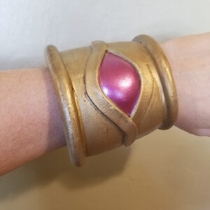 Pode incluir: Uma pulseira de punho dourada com uma pedra rosa iridescente em forma oval no centro. A pulseira tem uma superf&iacute;cie texturizada e um design que parece ser inspirado em temas antigos ou de fantasia. A pulseira &eacute; usada no pulso.