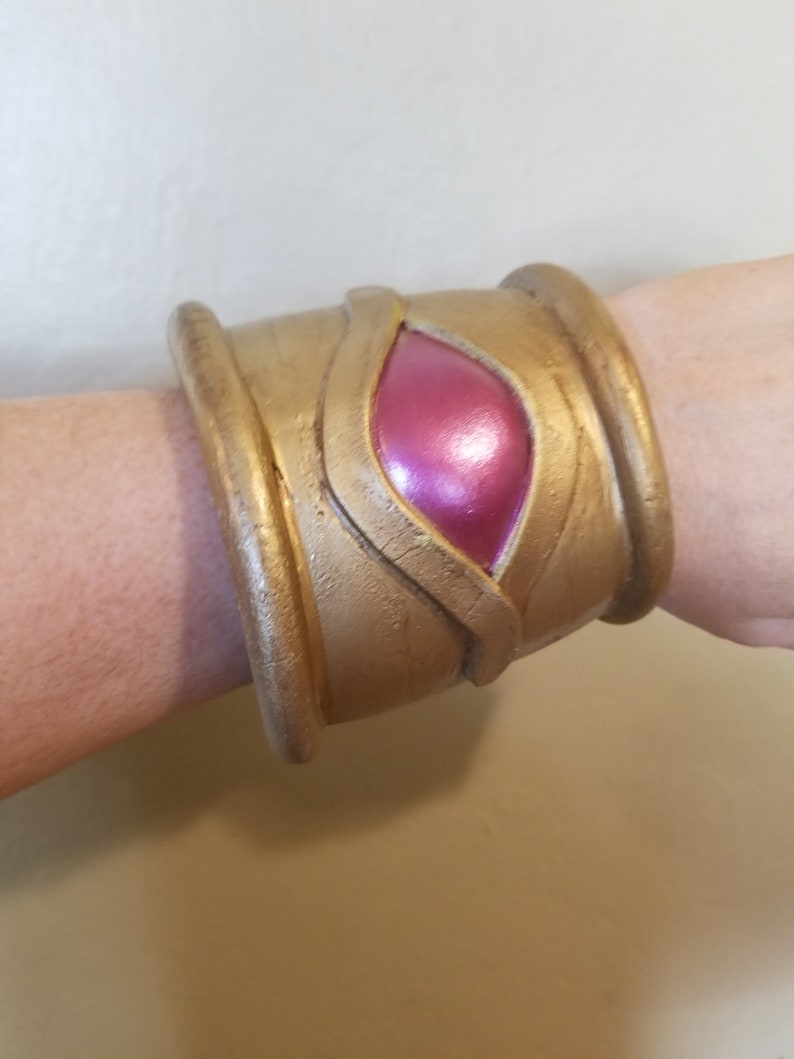 Pode incluir: Uma pulseira de punho dourada com uma pedra rosa iridescente em forma oval no centro. A pulseira tem uma superf&iacute;cie texturizada e um design que parece ser inspirado em temas antigos ou de fantasia. A pulseira &eacute; usada no pulso.