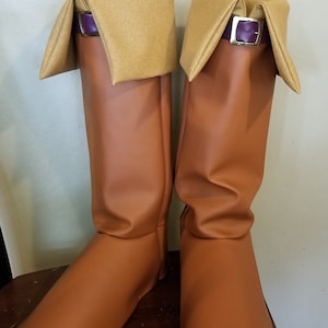 Puede incluir: Un par de botas marrones hasta la rodilla con punta cuadrada. Las botas tienen un pu&ntilde;o doblado en un color beige m&aacute;s claro, asegurado con una correa morada y una hebilla plateada. Las botas est&aacute;n hechas de un material liso similar al cuero.