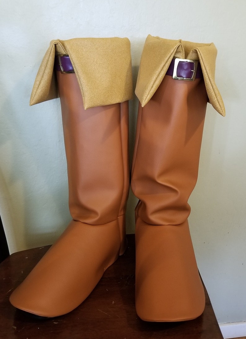 Puede incluir: Un par de botas marrones hasta la rodilla con punta cuadrada. Las botas tienen un pu&ntilde;o doblado en un color beige m&aacute;s claro, asegurado con una correa morada y una hebilla plateada. Las botas est&aacute;n hechas de un material liso similar al cuero.