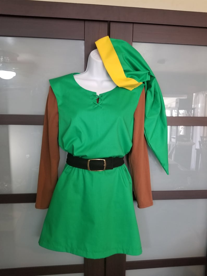 Pode incluir: Um traje de t&uacute;nica verde com mangas marrons, um cinto preto com fivela dourada e um chap&eacute;u verde combinando com um detalhe amarelo. O traje &eacute; exibido em um manequim, sugerindo um cosplay ou uma roupa tem&aacute;tica.