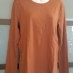 Pode incluir: Uma camisa de manga comprida com gola redonda em cor laranja queimado lisa. A camisa &eacute; exibida em um manequim branco. O tecido parece ser um material de malha macia. O fundo &eacute; de cor neutra.
