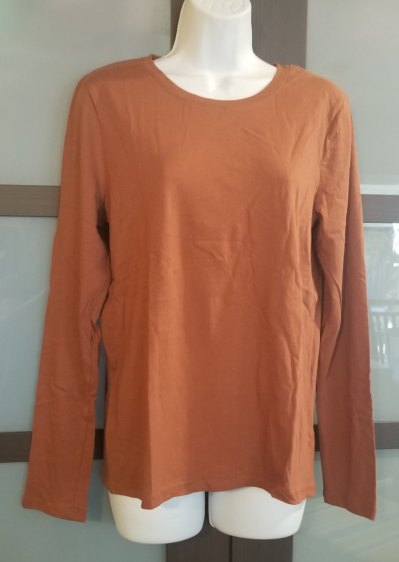 Pode incluir: Uma camisa de manga comprida com gola redonda em cor laranja queimado lisa. A camisa &eacute; exibida em um manequim branco. O tecido parece ser um material de malha macia. O fundo &eacute; de cor neutra.
