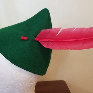 May include: A green felt hat with a triangular shape, featuring a vibrant red feather. The feather is inserted through the hat and secured with a small red accent. The hat is displayed on a white head form.