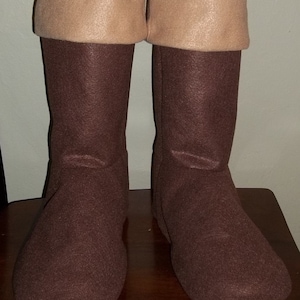 Pode incluir: Um par de botas de feltro castanhas com punhos bege. As botas, altas at&eacute; ao joelho, parecem feitas &agrave; m&atilde;o. Est&atilde;o sobre uma superf&iacute;cie de madeira.