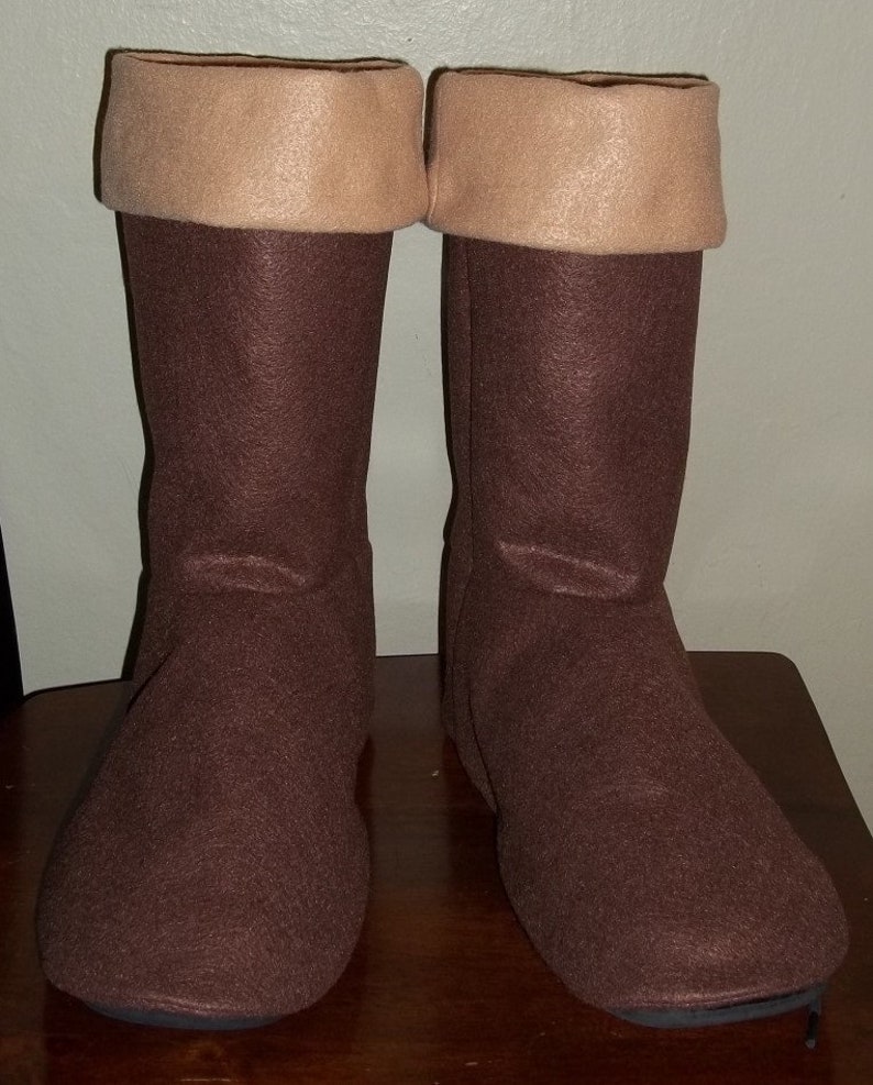 Pode incluir: Um par de botas de feltro castanhas com punhos bege. As botas, altas at&eacute; ao joelho, parecem feitas &agrave; m&atilde;o. Est&atilde;o sobre uma superf&iacute;cie de madeira.