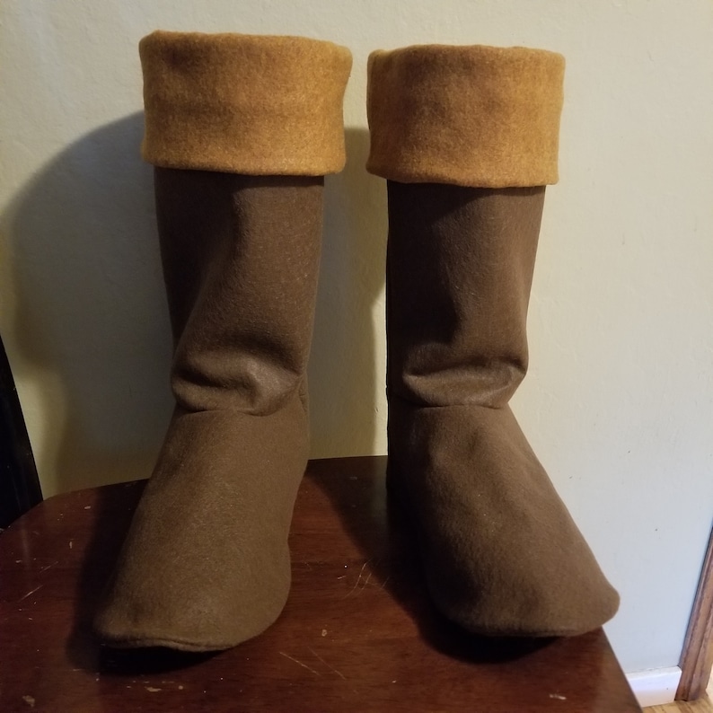 Pode incluir: Um par de botas de feltro marrom com uma dobra bege no topo. As botas v&atilde;o at&eacute; o joelho e parecem ser feitas &agrave; m&atilde;o. As botas est&atilde;o em uma superf&iacute;cie de madeira.