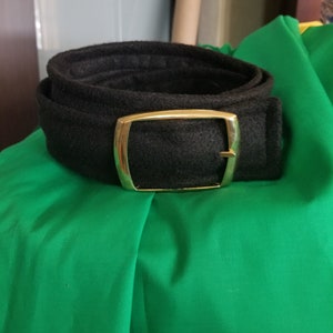 Pode incluir: Um cinto preto com fivela dourada enrolado sobre tecido verde. O cinto &eacute; feito de material texturizado. A fivela &eacute; retangular com cantos arredondados.
