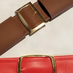 Puede incluir: Dos cinturones con hebillas rectangulares doradas. Uno es marr&oacute;n y el otro rojo. Ambos tienen costuras decorativas. El cintur&oacute;n marr&oacute;n est&aacute; abrochado, el rojo desabrochado. Adecuado para pantalones o faldas.