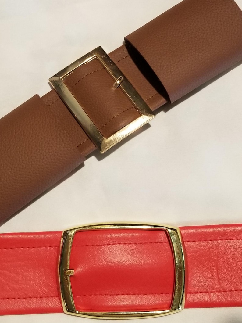 Puede incluir: Dos cinturones con hebillas rectangulares doradas. Uno es marr&oacute;n y el otro rojo. Ambos tienen costuras decorativas. El cintur&oacute;n marr&oacute;n est&aacute; abrochado, el rojo desabrochado. Adecuado para pantalones o faldas.